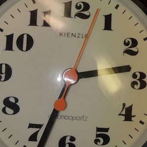 May include: A round Kienzle monoquartz wall clock with a cream-coloured face and large black serif numerals from 1 to 12. The clock features black hour and minute hands, a thin orange second hand, and the text "MADE IN GERMANY" at the bottom.