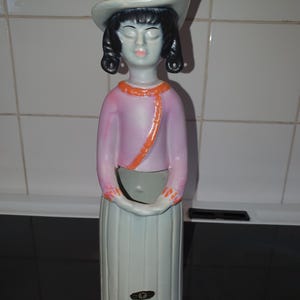 Puede incluir: Figura de cerámica vintage de una mujer con sombrero, top rosa con ribete naranja y falda blanca. La figura tiene cabello negro y ojos cerrados. La base es de color gris claro.