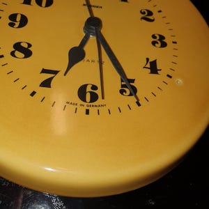 May include: A round yellow wall clock with a black face featuring bold black Arabic numerals from 1 to 12. The clock has black hour, minute, and second hands. The text "SINGER", "QUARTZ", and "MADE IN GERMANY" is printed in black on the clock face.