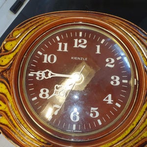 May include: A round Kienzle wall clock featuring a dark brown face with white Arabic numerals and ornate white hands. The clock is encased in a thick, textured ceramic frame with a glossy brown finish and decorative yellow flame-like accents.