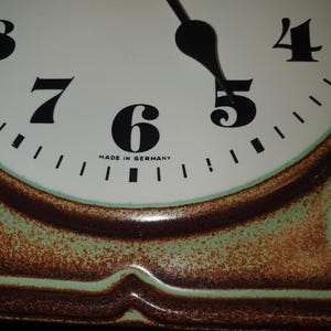 Peut inclure: Un cadran d'horloge avec un fond blanc, des chiffres noirs et des aiguilles noires. Le texte MADE IN GERMANY est imprimé sous le chiffre 6. L'horloge présente une bordure décorative avec un centre vert clair et un bord brun texturé.