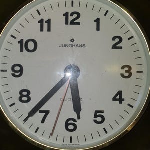 Puede incluir: Un reloj de pared redondo con esfera blanca, números arábigos negros y un borde dorado. El reloj tiene manecillas negras para las horas y los minutos, un segundero rojo fino y el texto JUNGHANS y QUARTZ. En la parte inferior dice MADE IN GERMANY.