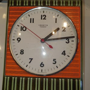 Peut inclure: Une horloge murale rectangulaire avec un cadran circulaire crème, des chiffres noirs et l'inscription VEDETTE transistor. Le cadre est orange avec des lignes horizontales et des bordures à rayures verticales vert olive foncé.