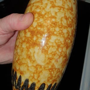 May include: A tall, oval ceramic vase with a speckled tan and brown pattern. The top and bottom of the vase feature a dark brown, zig-zag design. The vase is held by a person's hand.