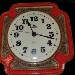 Puede incluir: Un reloj de pared cuadrado con esfera color crema, números negros y manecillas negras. El reloj presenta el texto "vedette" y "quartz" en la esfera. El marco es de cerámica con un borde rojo brillante y detalles en marrón moteado.