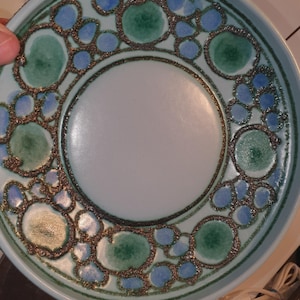 Pode incluir: Um prato redondo de cerâmica com um centro azul claro e uma borda decorativa. A borda apresenta um padrão de círculos verdes e azuis delineados em uma cor marrom escuro. O prato tem uma borda ligeiramente elevada.