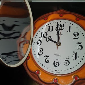 Puede incluir: Un reloj de pared redondo con esfera blanca, números negros y manecillas negras ornamentadas. El marco es naranja con detalles circulares rojos y un espejo circular abatible unido al lado izquierdo. La esfera muestra el texto AUNA y QUARTZ.