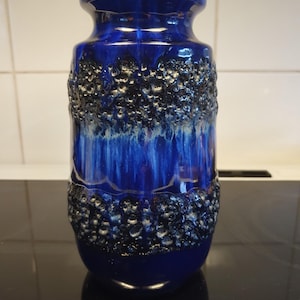 Puede incluir: Un jarrón de cerámica azul cobalto con una banda texturizada gris oscuro en el centro. El jarrón tiene un acabado brillante y una forma única. La parte superior e inferior son de color azul liso. Objeto decorativo.