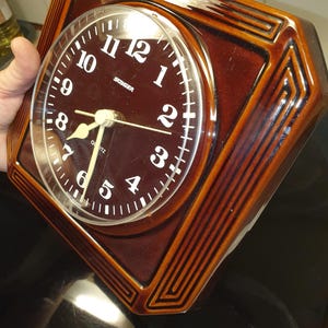 May include: A brown ceramic wall clock with a glossy finish and a geometric border. The round clock face features white Arabic numerals, white hands, and the text Stager and Quartz printed in white. It includes the text West Germany at the bottom.