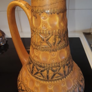 Op de afbeelding: Een hoge keramische kan met een warme oranje en bruine glazuurlaag. Het oppervlak is voorzien van een herhalend patroon van reliëfcirkels en donkere, ingesneden geometrische kruisarceringen. De kan heeft één gebogen handvat aan de linkerzijde.