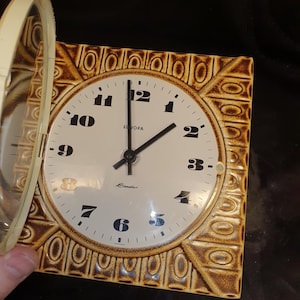 Puede incluir: Un reloj de pared cuadrado y vintage con esfera crema y números negros. El reloj tiene un borde marrón texturizado. El reloj marca las 2:00. La palabra "Europa" está impresa en la esfera.