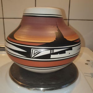 Könnte beinhalten: Eine handbemalte Keramikvase mit rundem Körper und breiter Öffnung. Die Vase zeigt eine Sonnenuntergangsszene mit einer schwarzen Silhouette einer Struktur. Das Design umfasst schwarz-weiße geometrische Muster und orangefarbene Bänder.