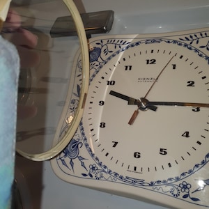 May include: A square wall clock with an off-white face and blue floral patterns along the edges. The clock features black numerals from 1 to 12, black hands, and the text Kienzle Automatic and Made in Germany. A circular glass cover is hinged open.
