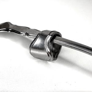 May include: A handcrafted, bent silverware art piece. The piece features a fork head bent around a spoon handle, creating a unique knot-like design. The silverware is made of shiny, polished metal, likely stainless steel.
