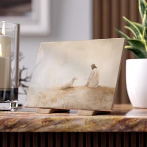 Könnte beinhalten: Ein gerahmtes Gemälde, das einen Mann in weißen Gewändern zeigt, der in einem Feld kniet, neben ihm steht ein weißes Lamm. Der Hintergrund ist in einem weichen, gedämpften Beige gehalten.