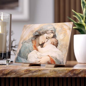 May include: A watercolour painting of the Virgin Mary holding a sleeping baby Jesus. The painting is on a wooden plaque with a stand.