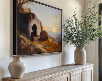 May include: A framed landscape painting depicts a figure in a dark robe approaching a cave entrance at sunset. The artwork is mounted on a cream-colored wall above a wooden cabinet. Two decorative vases and greenery complete the scene.