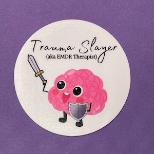 May include: A white circle sticker with a pink brain cartoon character holding a sword and a shield. The text on the sticker reads "Trauma Slayer (aka EMDR Therapist)"