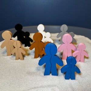 May include: A collection of wooden figure cutouts in various colors, including tan, black, white, brown, gray, pink, and blue. Each figure is a stylized representation of a person, with a larger figure paired with a smaller one.