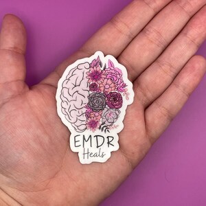 May include: A white sticker with a pink and purple floral design inside a brain outline. The text "EMDR Heals" is below the brain.