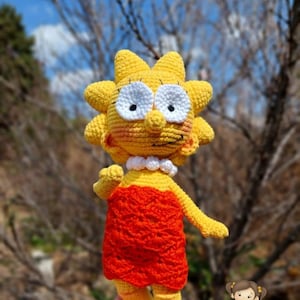 May include: A crocheted doll of Lisa Simpson from The Simpsons. She is wearing a red dress and yellow shoes. The doll is being held in a person's hand.