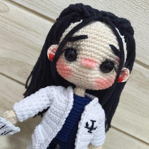 May include: Crocheted doll depicting a doctor, with black hair and large eyes. The doll wears a white coat over a navy blue outfit, with a medical symbol on the coat. The doll's face has rosy cheeks and red accents on the ears.