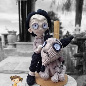 May include: A crocheted doll with black hair and a gray and white dog. The doll is wearing black pants and a gray shirt. The dog is sitting on a wooden stand. The doll is holding the dog's back. The background is blurred.
