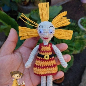May include: Crocheted doll with yellow hair, a red and yellow striped dress, and a black belt with a yellow buckle. The doll is wearing red shoes.