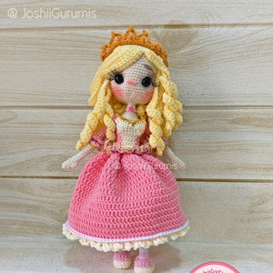 Może przedstawiać: Lalka szydełkowa w różowej sukience z białą koronkową lamówką, złotą koroną i blond włosami. Lalka stoi na drewnianej powierzchni.