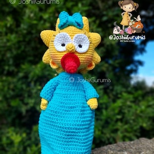 May include: A crocheted yellow doll with a blue bow, blue eyes, and a red pacifier. The doll is wearing a blue crocheted dress and is standing on a wooden base. The doll is a handmade creation by @JoshiiGurumis.