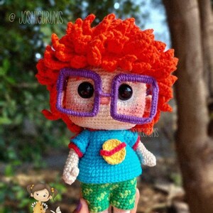 Peut inclure: Une poupée amigurumi en crochet avec des cheveux roux, des lunettes violettes, un t-shirt bleu avec un motif de planète jaune, un short vert et des chaussures rouges. La poupée tient un petit animal en crochet dans sa main.