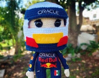 May include: Crocheted amigurumi doll of a race car driver wearing a blue and red racing suit with the Oracle and Red Bull logos. The doll is wearing a white helmet with a blue visor and a yellow stripe. The doll is holding a Mexican flag in its right hand.