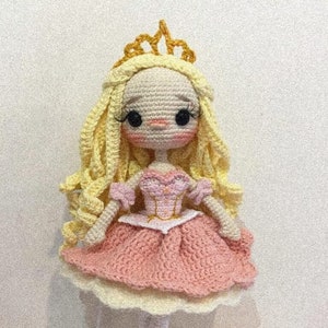 May include: A crocheted doll wearing a pink dress and a gold crown. The doll has blonde hair and large, black eyes.