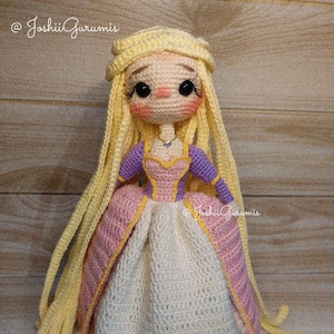 May include: A crocheted doll with long blonde hair, wearing a purple and pink dress with a white underskirt. The doll has a light pink face with black eyes and a light blue necklace. The doll is standing on a wooden surface. @JoshiiGurumis