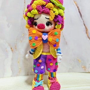 May include: A handmade crochet clown doll with a colorful design. The doll features a pink hat, a yellow and green wig, and a large orange bow tie with colorful polka dots. The clown wears a yellow jacket, purple and pink polka dot pants, and rainbow striped socks and shoes.