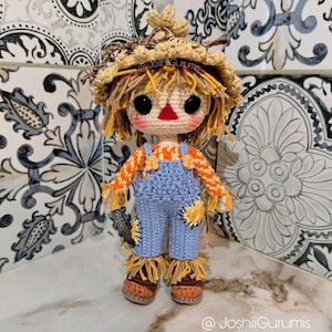 May include: A crocheted scarecrow doll with a straw hat, blue overalls, and a yellow and orange shirt. The doll is standing on a white tiled surface.