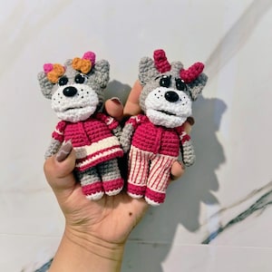 May include: Two handmade crochet dog dolls. The dogs have gray heads, white faces, and black eyes. They wear pink outfits with white and red accents and bows. The dolls are held in a hand.