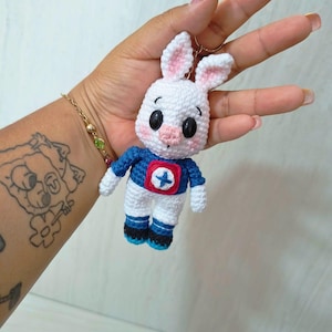 May include: A handmade white crochet bunny keychain with pink inner ears, black eyes, and a pink nose. The bunny wears a blue and red superhero-style outfit with a white star. The keychain is a cute, whimsical accessory.