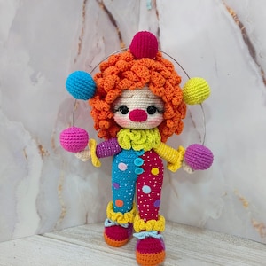 May include: A handmade crochet clown doll with orange hair, a red nose, and colorful clothing. The doll has blue and red polka-dot pants, a yellow collar, and is holding colorful balls. The clown's hair is topped with a red ball.