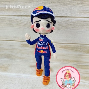 May include: Crocheted amigurumi doll wearing a blue racing suit with red accents. The suit has the words "Oracle" and "Red Bull" on it. The doll is wearing a red and white racing cap and has a friendly expression.