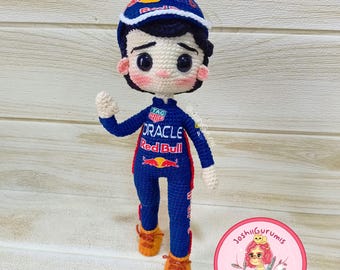 May include: Crocheted amigurumi doll wearing a blue racing suit with red accents. The suit has the words "Oracle" and "Red Bull" on it. The doll is wearing a red and white racing cap and has a friendly expression.