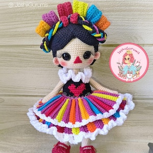 May include: A colorful crocheted doll wearing a multi-colored dress with a white ruffle. The doll has black hair with a braid and a red heart on her dress. The doll is sitting on a wooden surface. @JoshiiGurumis