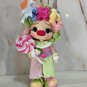 May include: A handmade crochet doll in the shape of a clown, holding a lollipop. The doll has a colorful, curly wig, a red nose, and a bow on its hat. The clown is wearing a patchwork outfit of pink, green, and yellow.