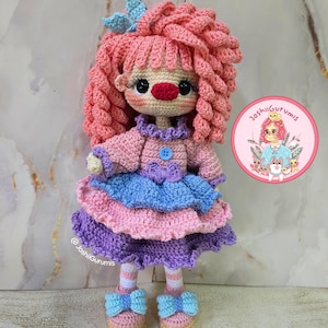 May include: A handmade crochet doll with pink curly hair, a red nose, and a multi-layered dress in shades of pink, blue, and purple. The doll has a blue bow on its head and wears shoes with blue bows. The doll is labeled with the text "@ JoshiiGrumis".