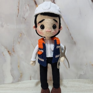 May include: Crocheted doll depicting a construction worker. The doll wears a white hard hat, an orange and navy blue vest, black pants, and brown boots. It holds a pair of silver pliers. The doll has a neutral expression.