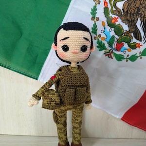 May include: A handmade crochet doll dressed in a military-style uniform, complete with a backpack and helmet. The doll has a neutral expression and is set against the backdrop of a Mexican flag, featuring green, white, and red colors.