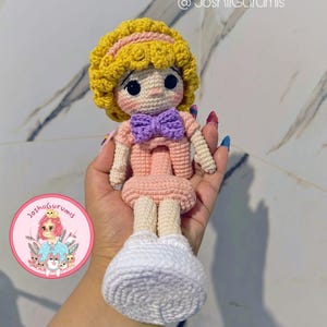 May include: A crocheted doll with yellow hair, a pink dress, and a purple bow. The doll is holding a white crocheted purse. The doll is being held by a hand with a tattoo.