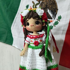 May include: A handmade crochet doll in a white dress with red and green trim, a floral crown, and braids. The doll is positioned in front of a Mexican flag.