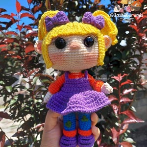 May include: A crocheted doll with yellow hair, purple bows, a purple dress, orange and blue trousers, and purple shoes. The doll is being held in a hand against a background of green leaves.