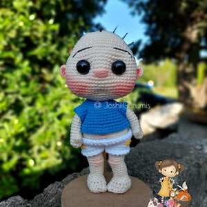 May include: A crocheted amigurumi doll of a boy wearing a blue shirt, white shorts, and a light brown base. The doll has a light brown complexion, black hair, and large black eyes. The doll is standing on a wooden base with the text "@joshiigurumis 100% hecho a mano" on it.
